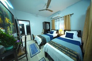 Habitación de hotel con dos camas, ventilador de techo, unidad de aire acondicionado, ventana con cortinas, un cuadro en la pared y una planta en maceta.