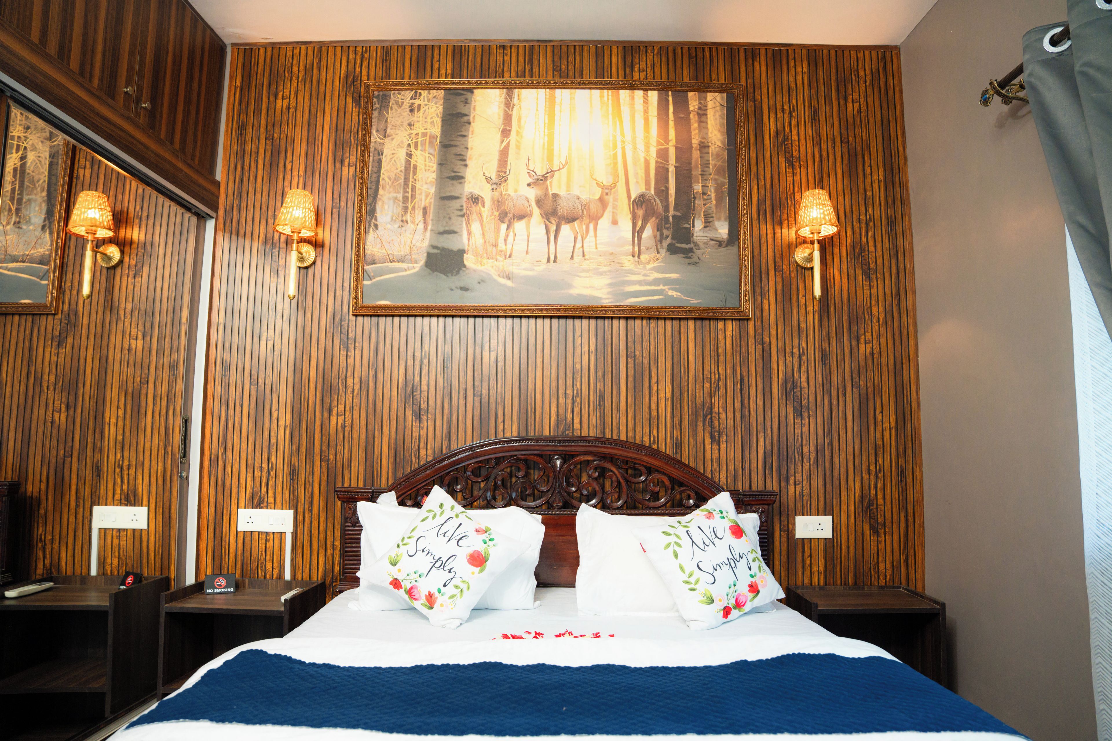 A hotel room with a wooden paneled wall, a large framed picture of deer in a forest, and two wall-mounted lamps.