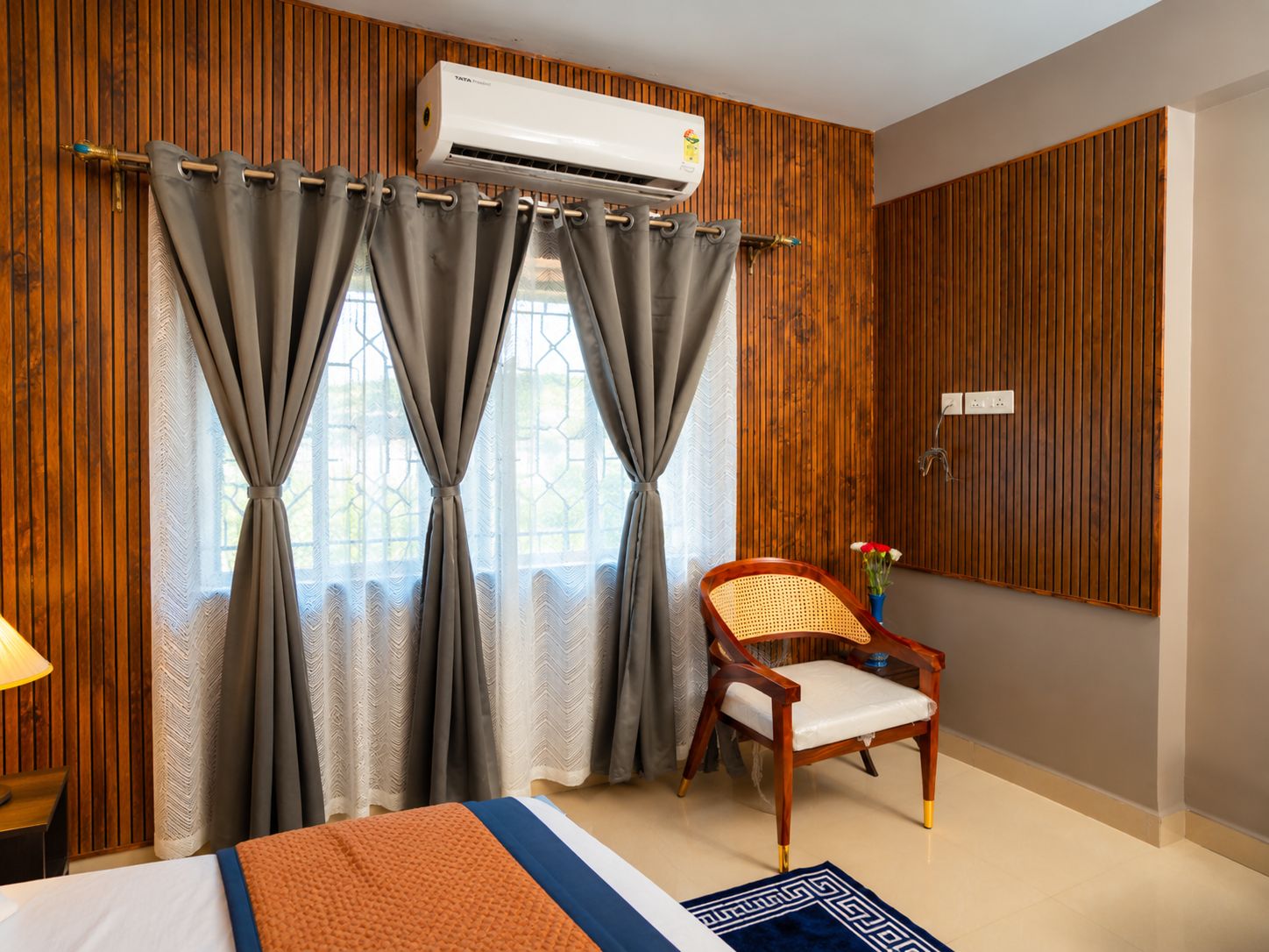 A hotel room with a bed, a chair, a wall-mounted air conditioner, and a window with curtains.