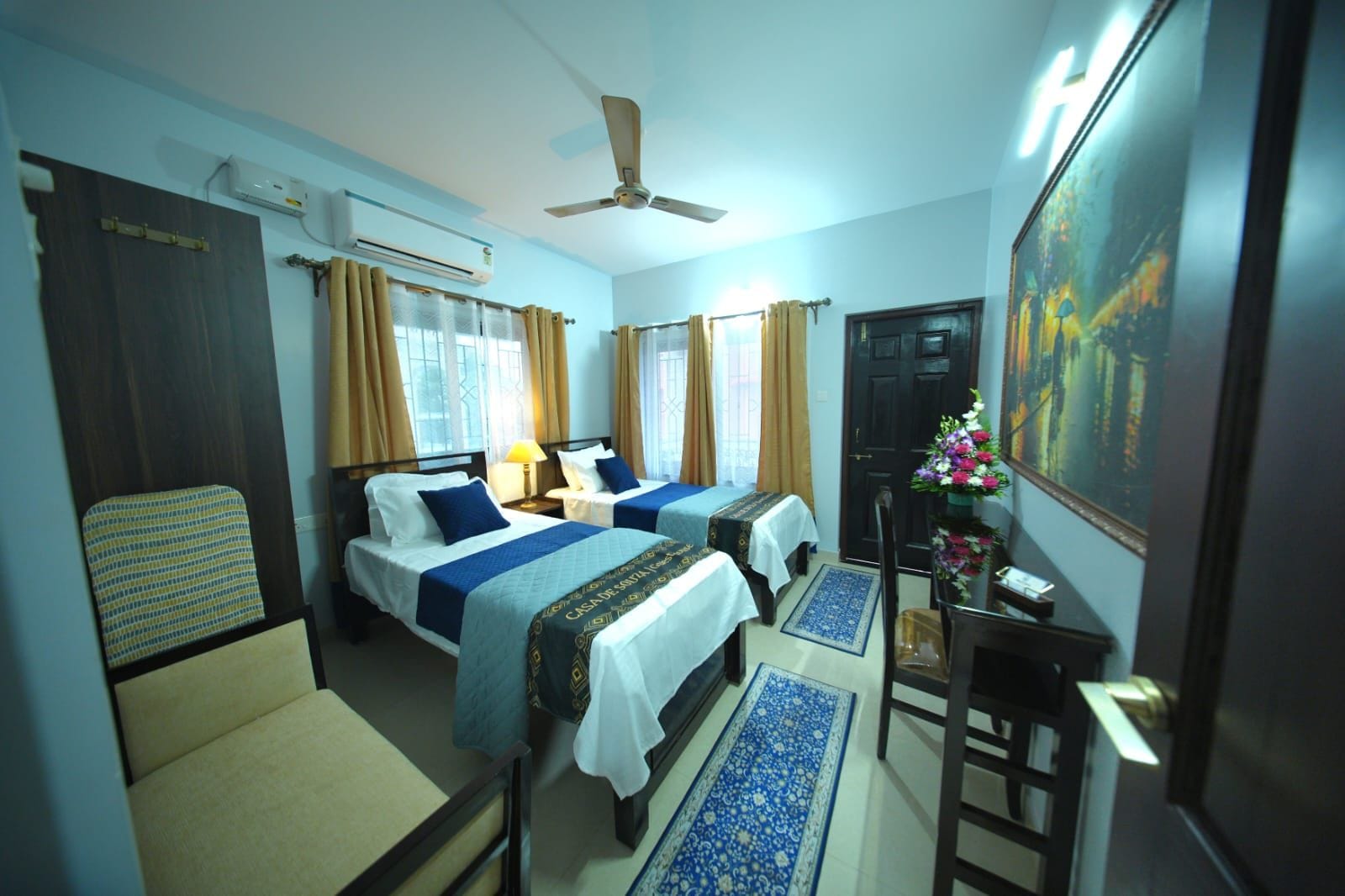 Une chambre d’hôtel avec deux lits, un bureau, une chaise, un ventilateur de plafond et un tableau au mur.