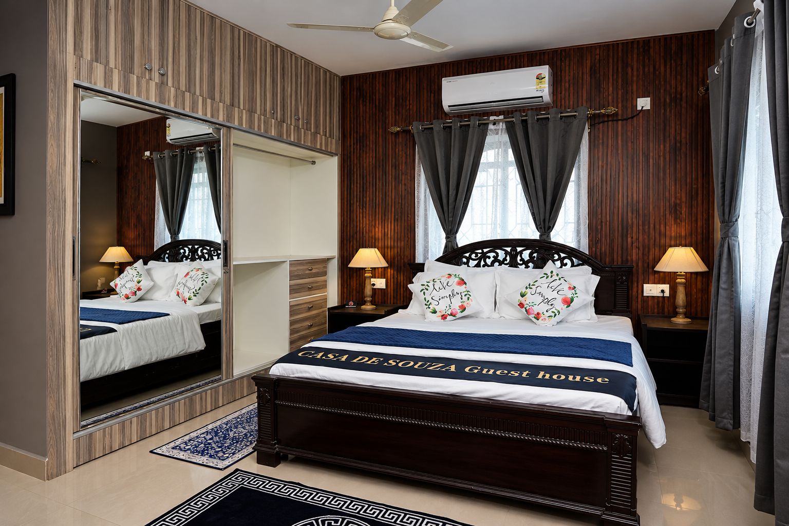 A guest room with a large bed, two bedside tables with lamps, a ceiling fan, an air conditioning unit, and a mirror.