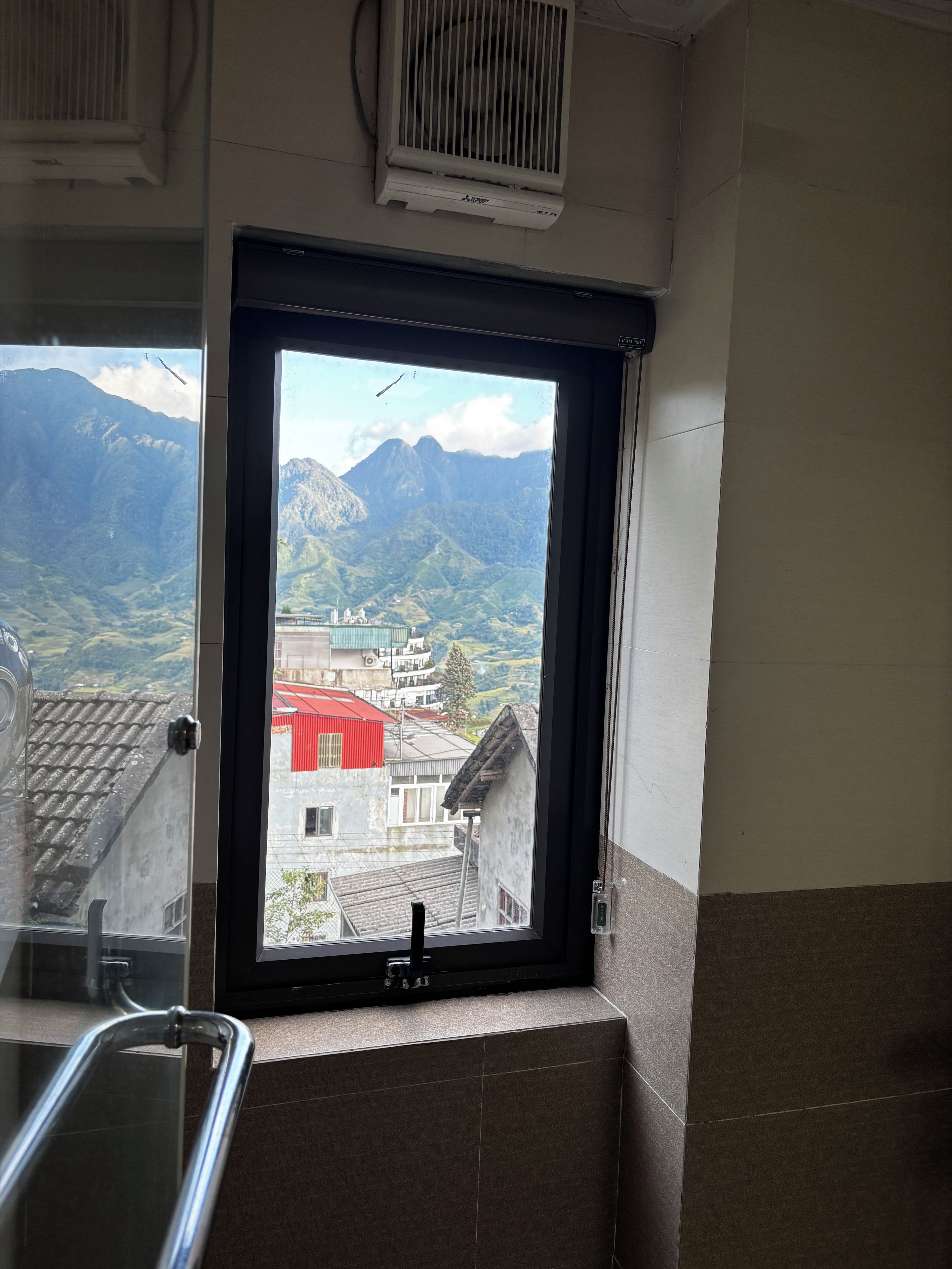 Un baño con ventana que ofrece vistas a un paisaje montañoso y un edificio de techo rojo.