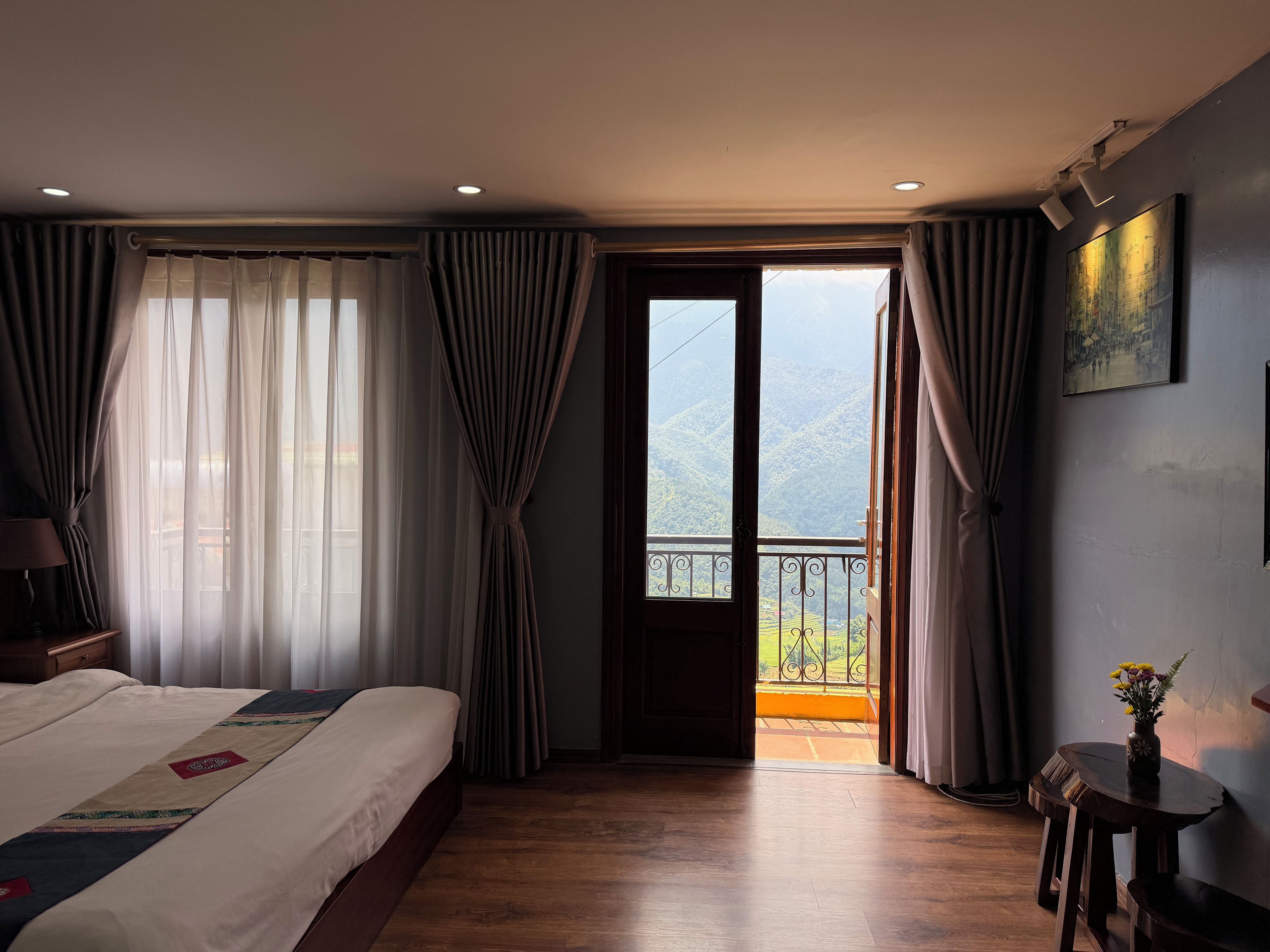 Habitación de hotel con cama, una mesa con un florero y un balcón con vistas a las montañas.