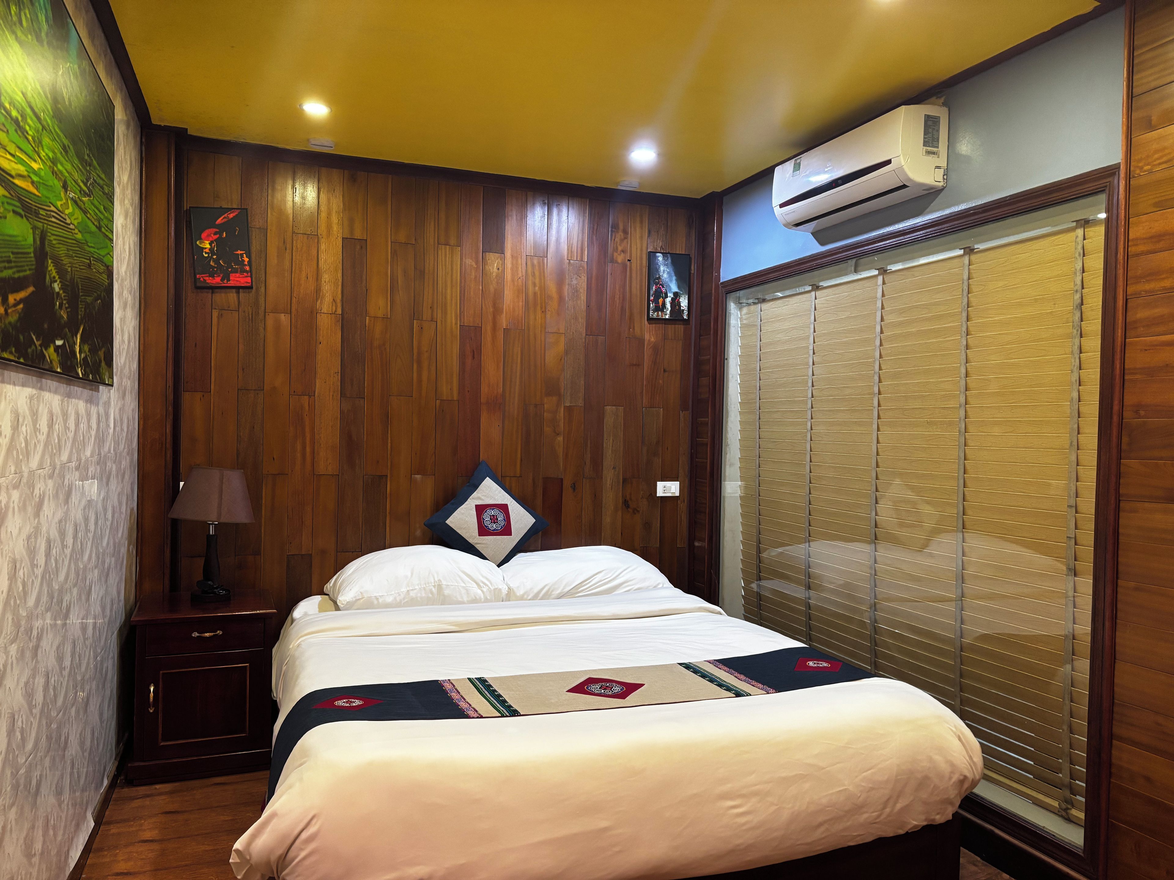 A hotel room with a wooden paneled wall, a bed with a decorative headboard, a nightstand with a lamp, and a window with blinds.