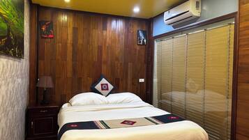 A hotel room with a wooden paneled wall, a bed with a decorative headboard, a nightstand with a lamp, and a window with blinds.