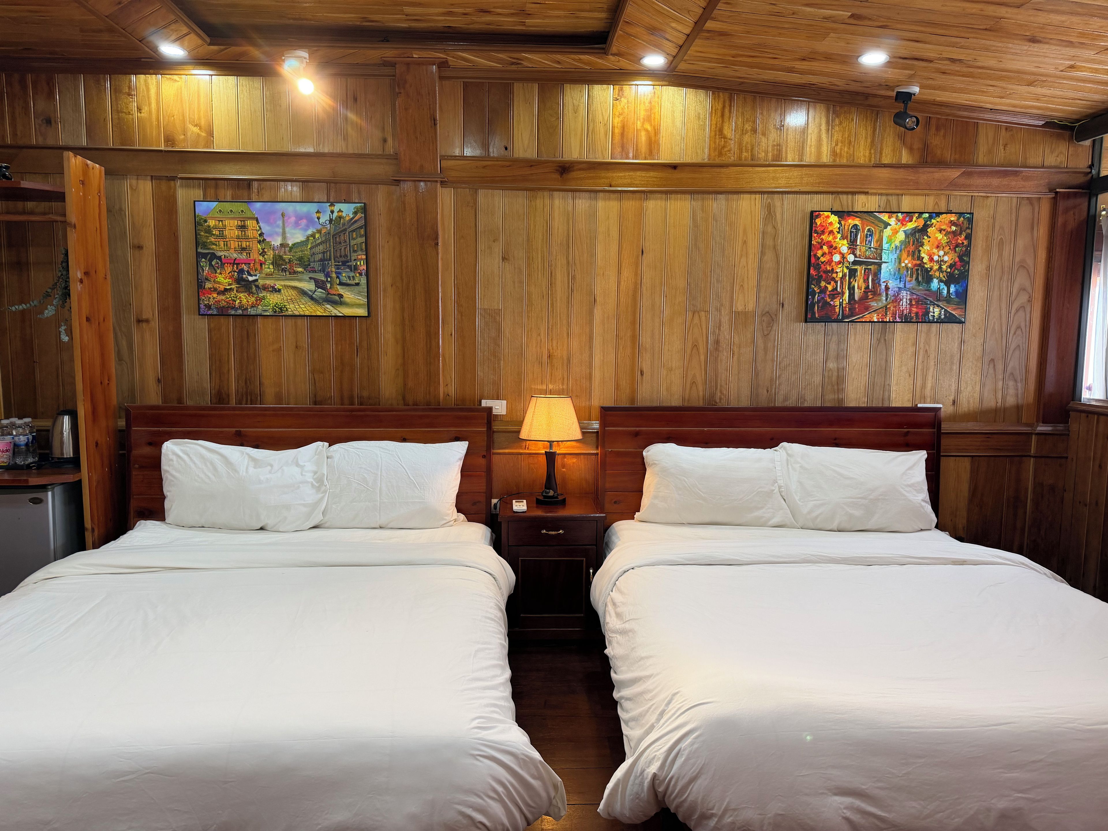 A room with two beds, wooden panel walls, and two paintings.