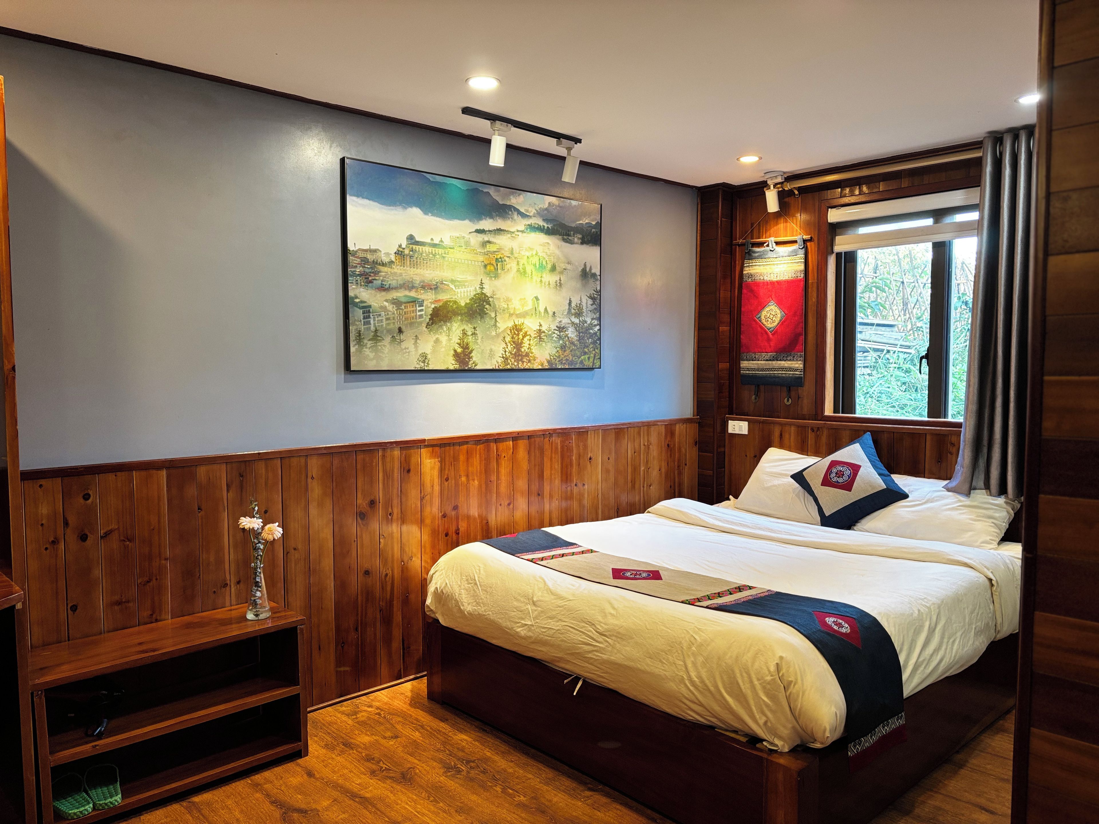 A hotel room with a bed, wooden paneling, a painting, a window with curtains, and a shelf with slippers.
