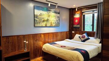 A hotel room with a bed, wooden paneling, a painting, a window with curtains, and a shelf with slippers.