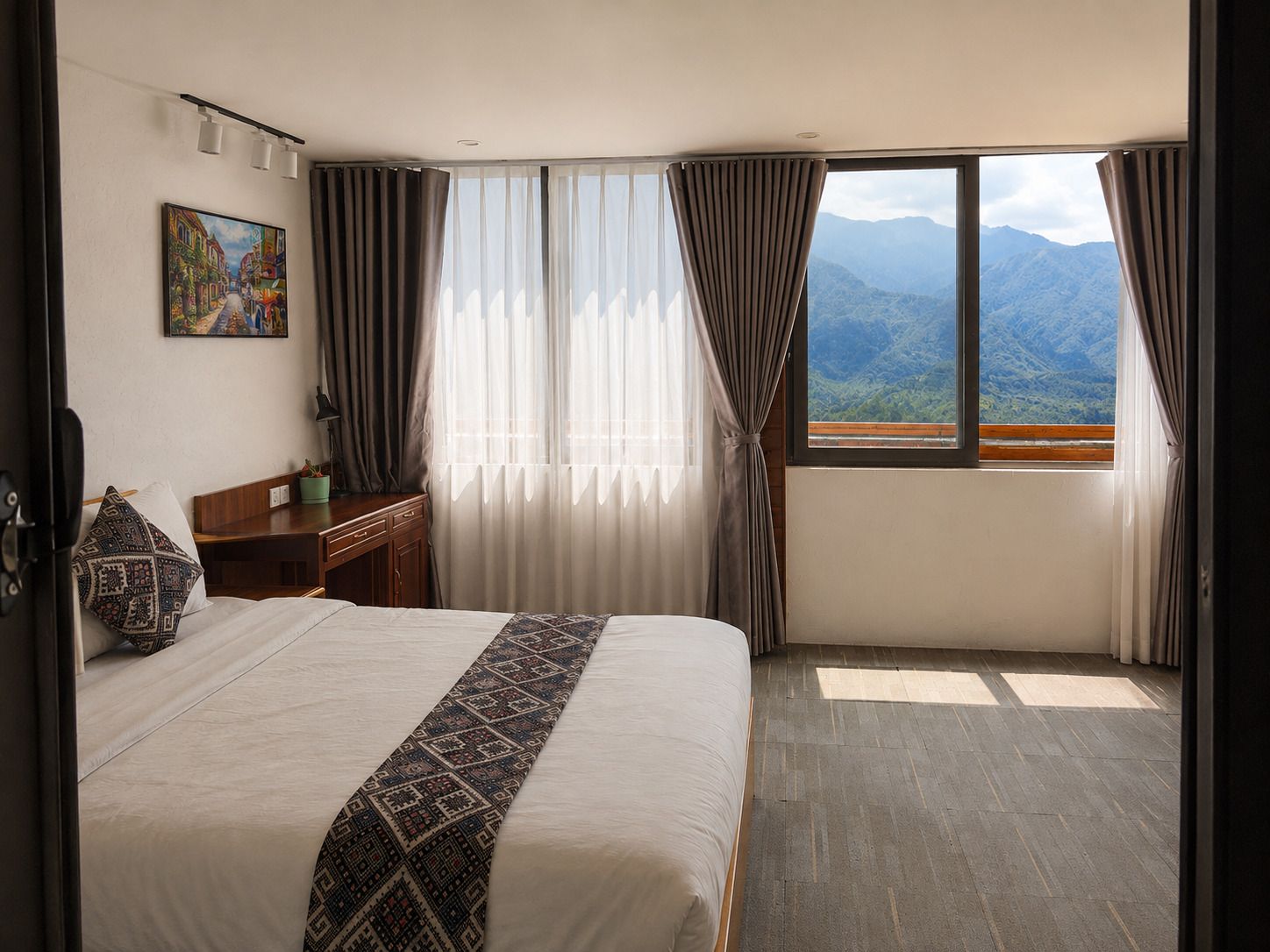 Habitación de hotel con una cama grande, un escritorio, una silla, un ventanal con vista a la montaña y un cuadro en la pared.