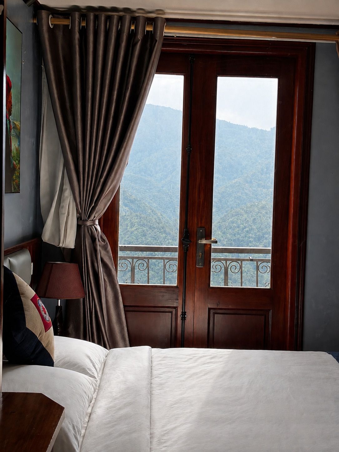 Ein Hotelzimmer mit Bett, Nachttisch, Lampe und Blick auf eine gebirgige Landschaft durch eine Tür.