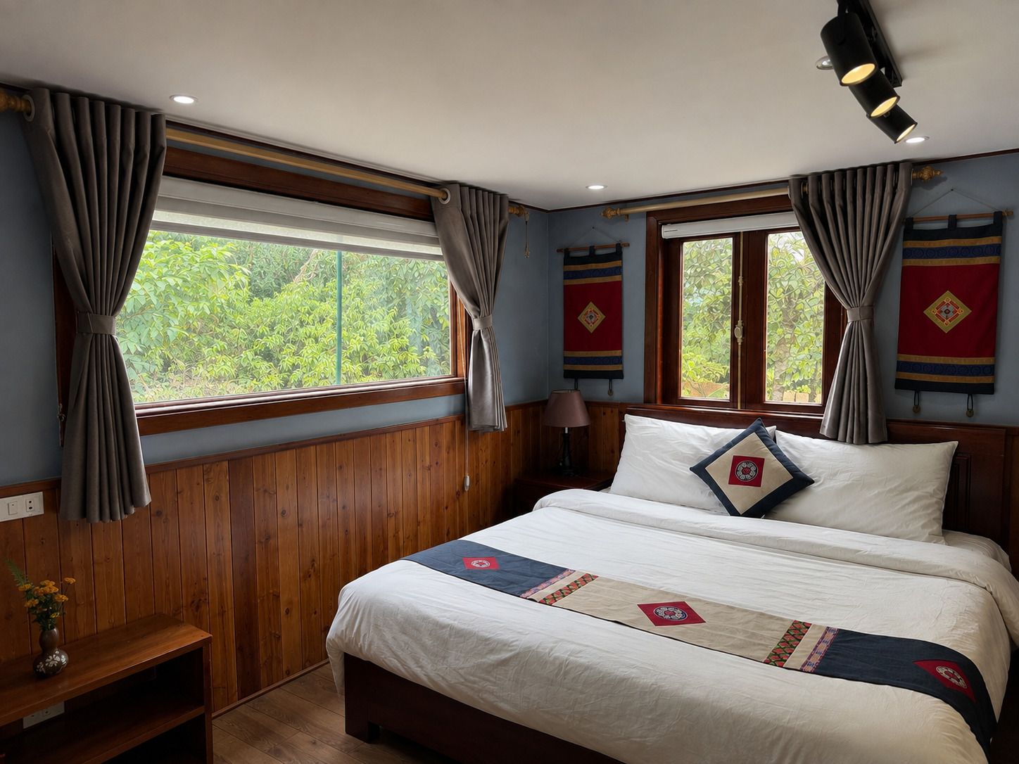 Un dormitorio con pared de paneles de madera, una cama con ropa de cama blanca y almohadones decorativos, una mesita de noche con lámpara y cortinas en las ventanas.