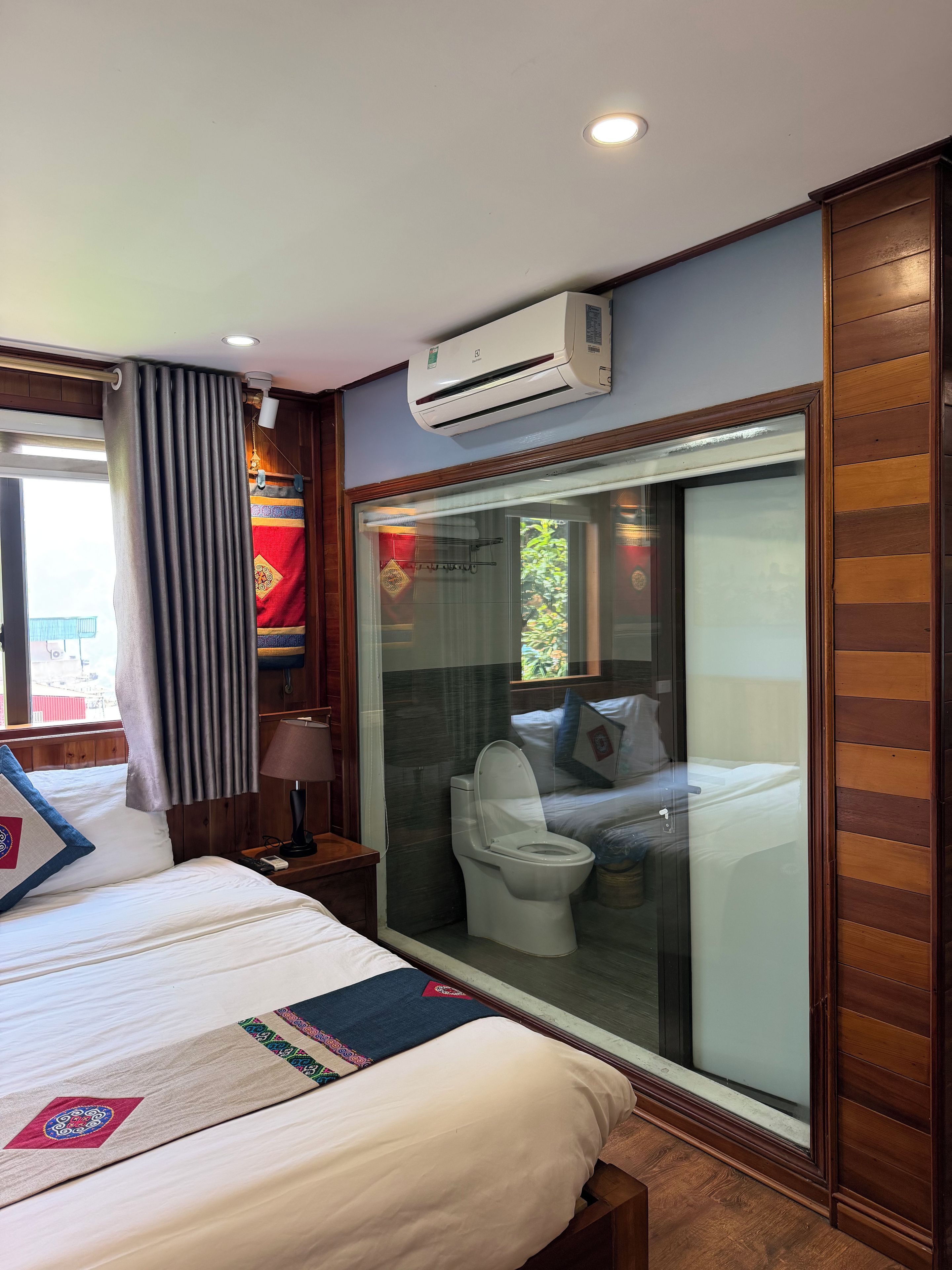 Habitación de hotel con cama, mesita de noche, lámpara, equipo de aire acondicionado y vistas al balcón a través de un ventanal.