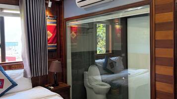 Habitación de hotel con cama, mesita de noche, lámpara, equipo de aire acondicionado y vistas al balcón a través de un ventanal.