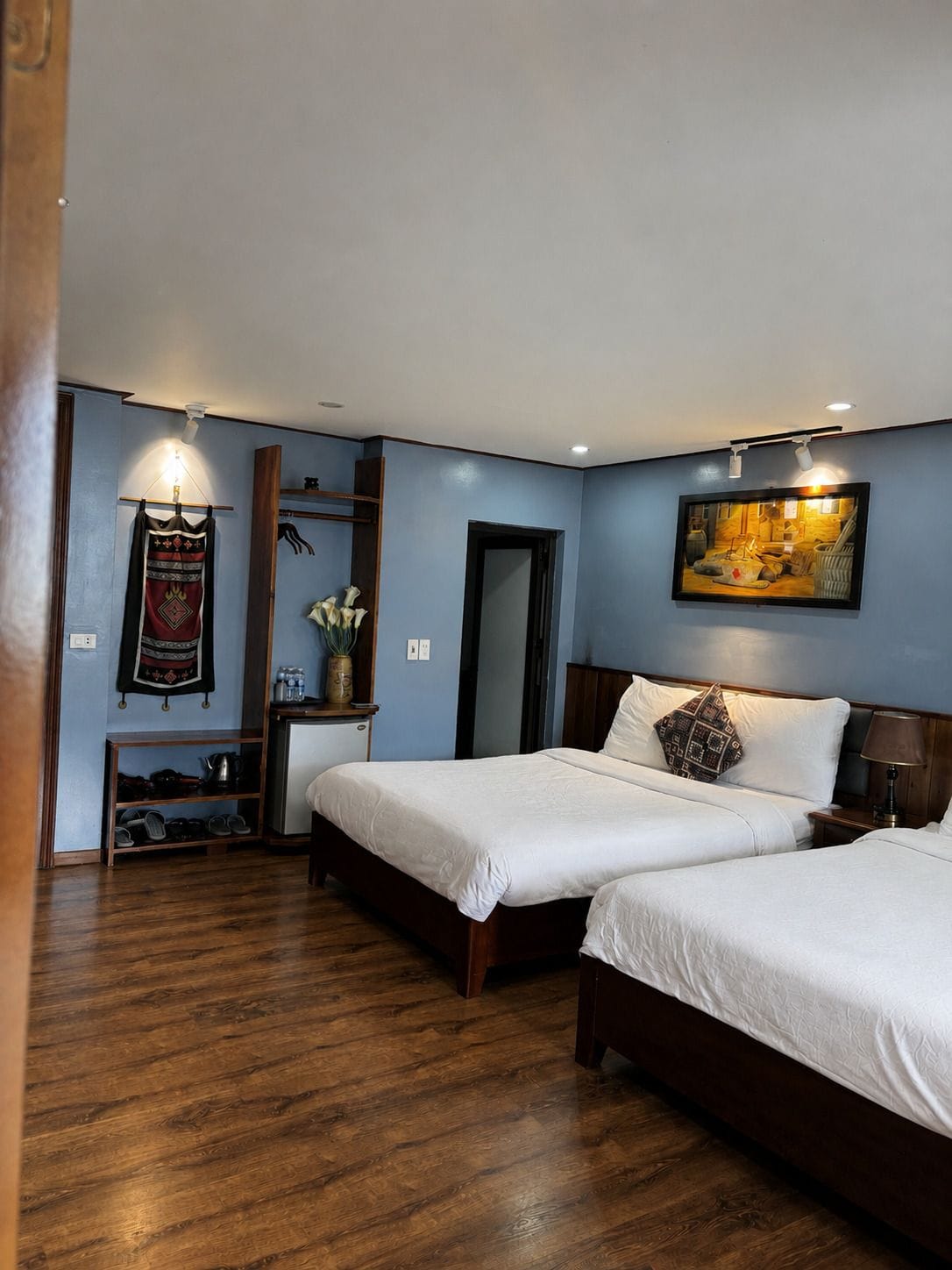 A hotel room with two beds, a wooden floor, a wall-mounted shelf with shoes, a painting, and a door.