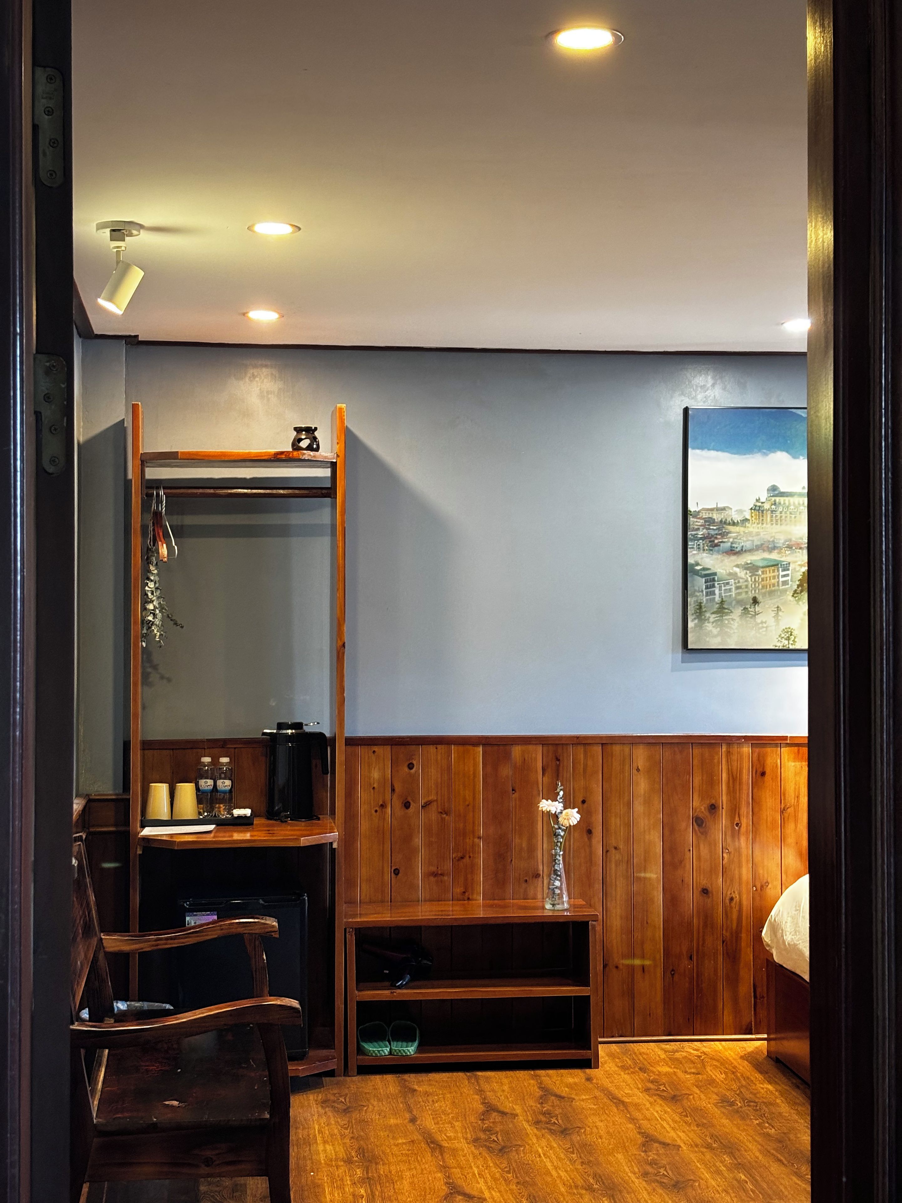 Una habitación con piso de madera, una cama, un escritorio con silla, un espejo, un estante con objetos y un cuadro en la pared.