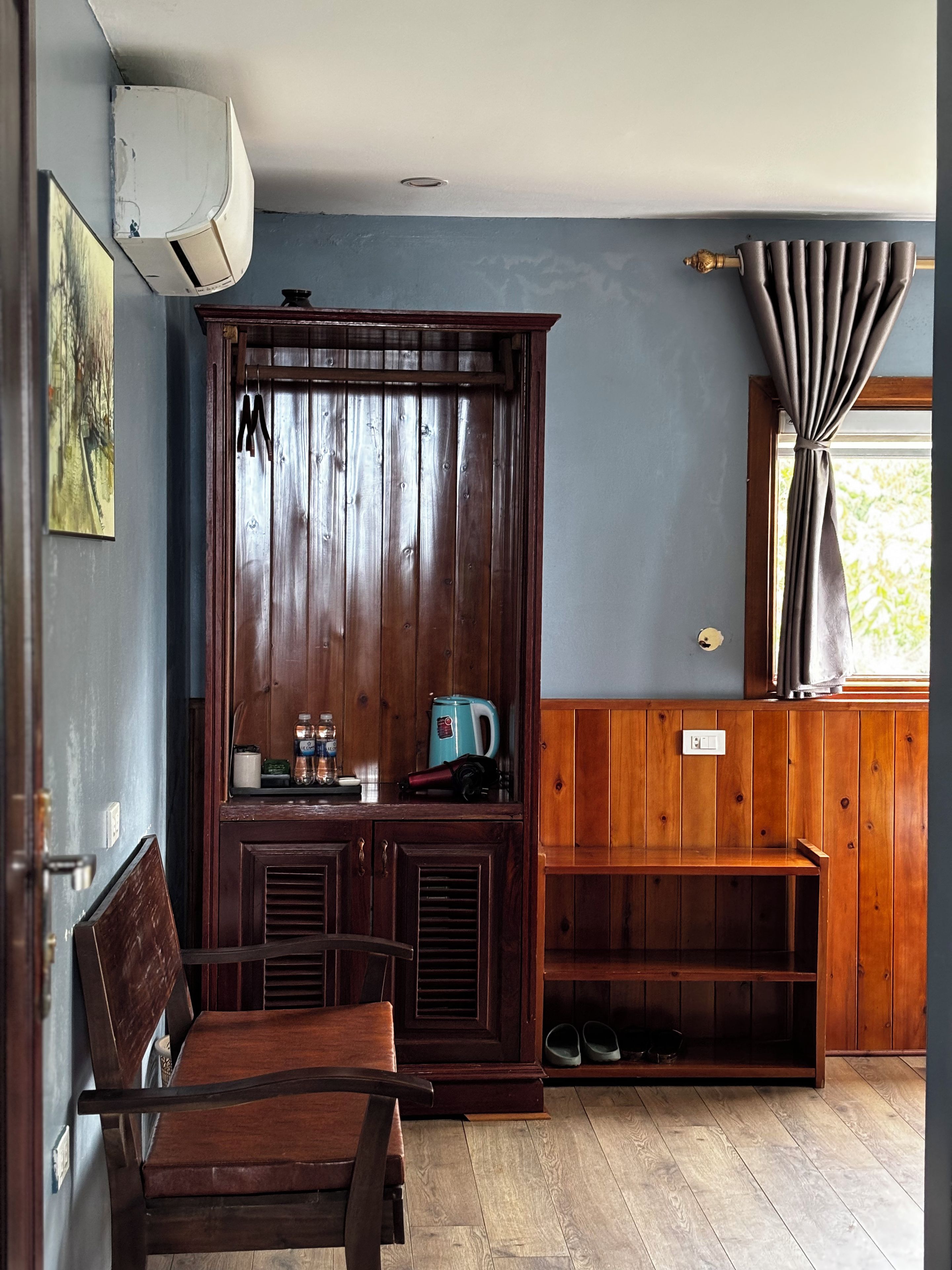 Una habitación con un armario de madera, un banco, una mesa y una ventana con cortinas.