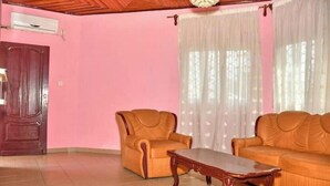 A room with a wooden ceiling, a brown door, a pink wall, a brown sofa, a wooden coffee table, and a window with sheer curtains. - NAL-Facing Apartment with Air Conditioning (Région du Littoral)