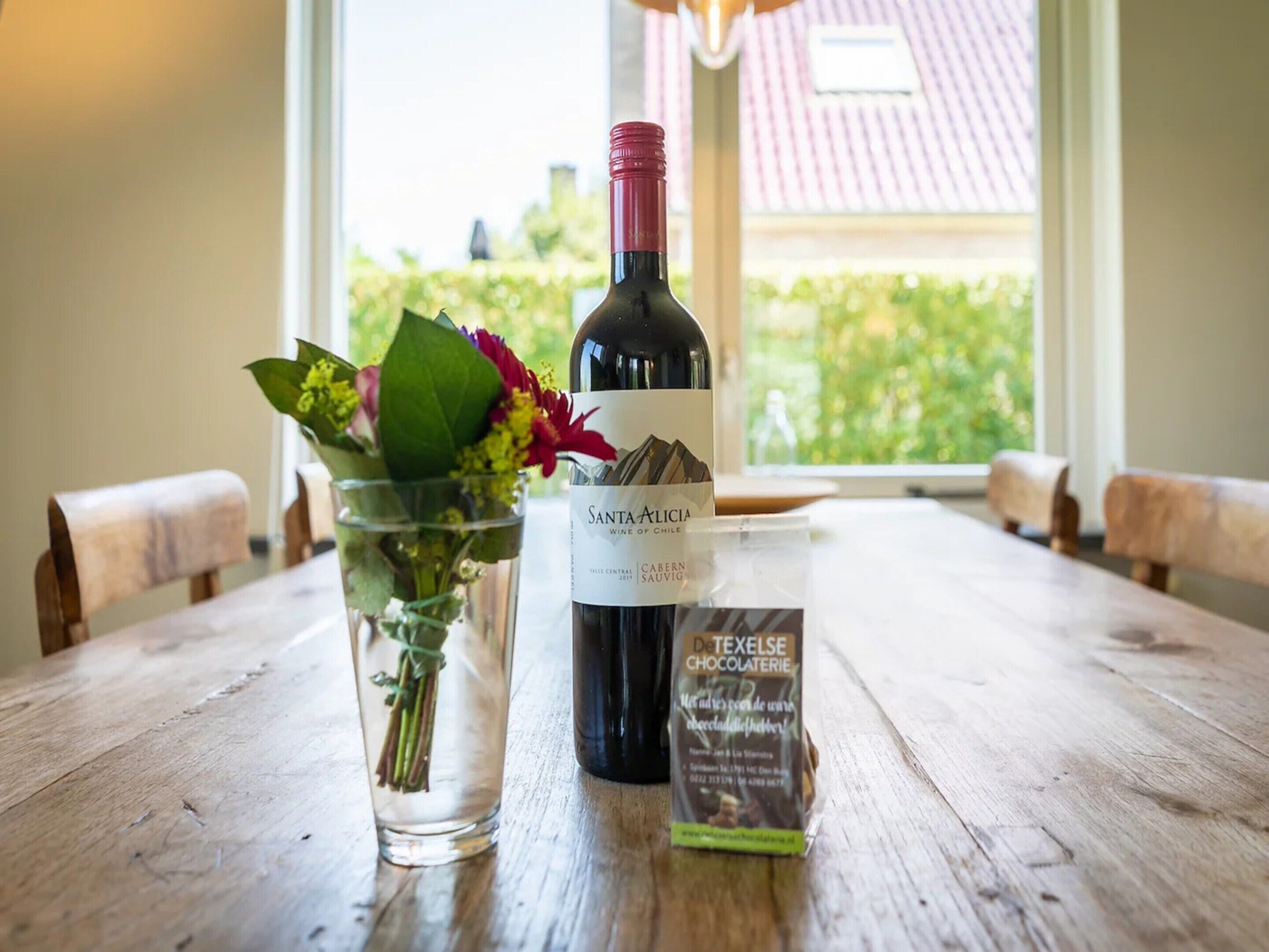 A bottle of wine, a glass with flowers, and a chocolate bar on a wooden table.