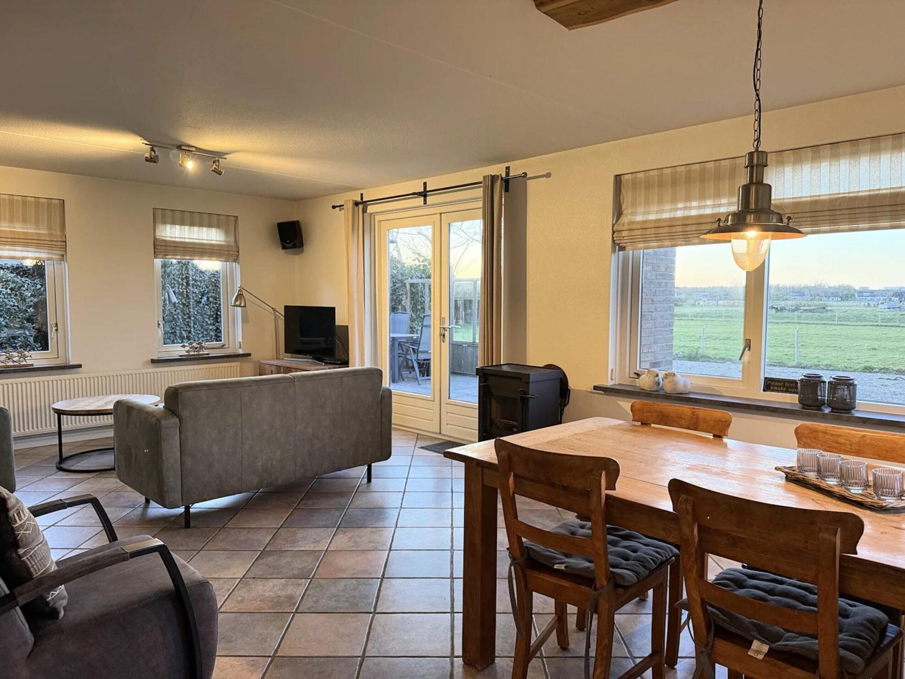 Ein geräumiger Wohnbereich mit einer Couch, einem Esstisch mit Stühlen, einem Fernseher und Blick ins Freie durch große Fenster.