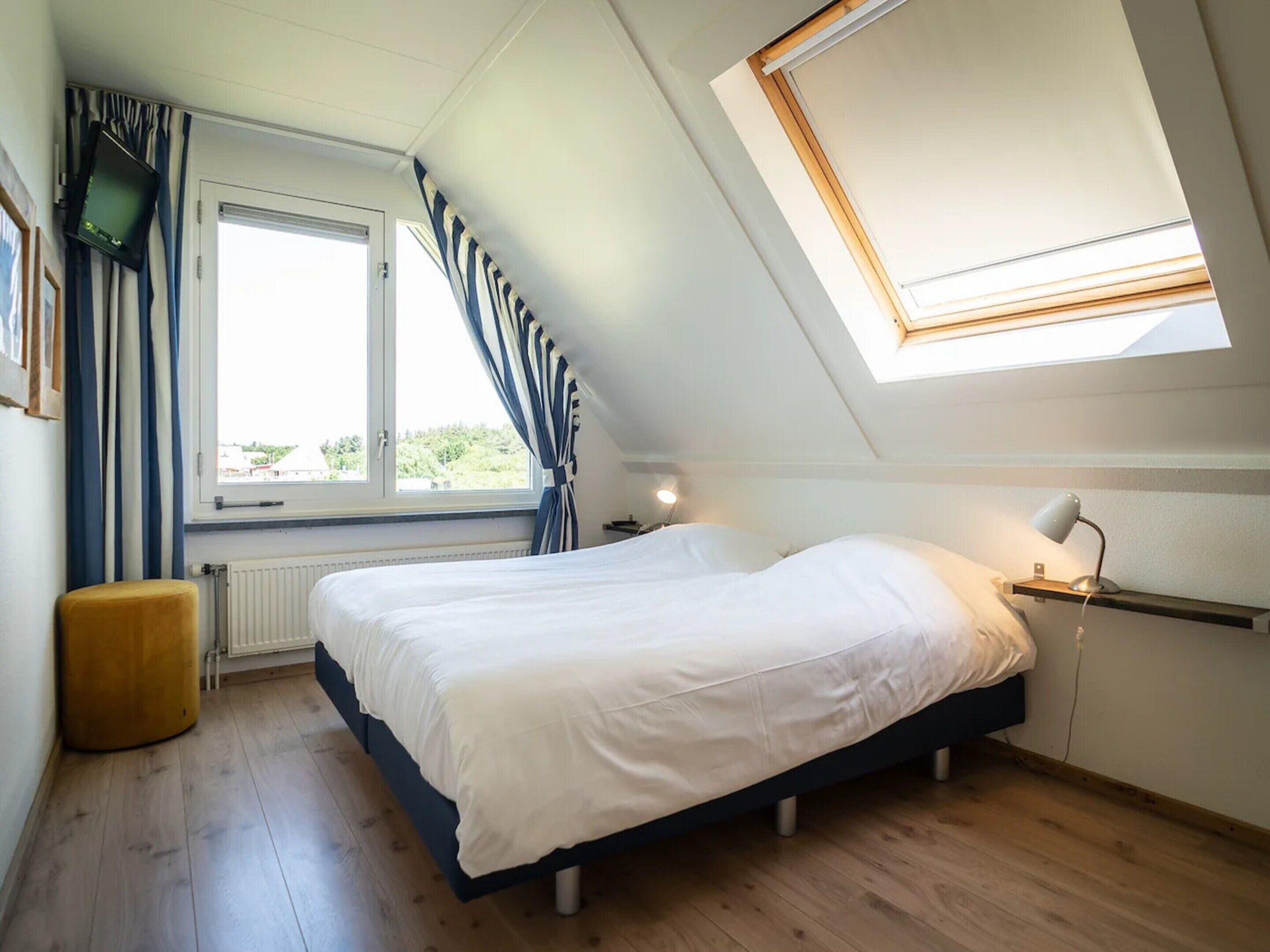 A bedroom with a bed, a window with curtains, a bedside table with a lamp, and a skylight.
