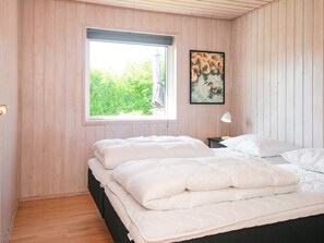 Une chambre avec une grande fenêtre, un lit recouvert de linge de lit blanc, une petite table de chevet avec une lampe et un tableau encadré accroché au mur.