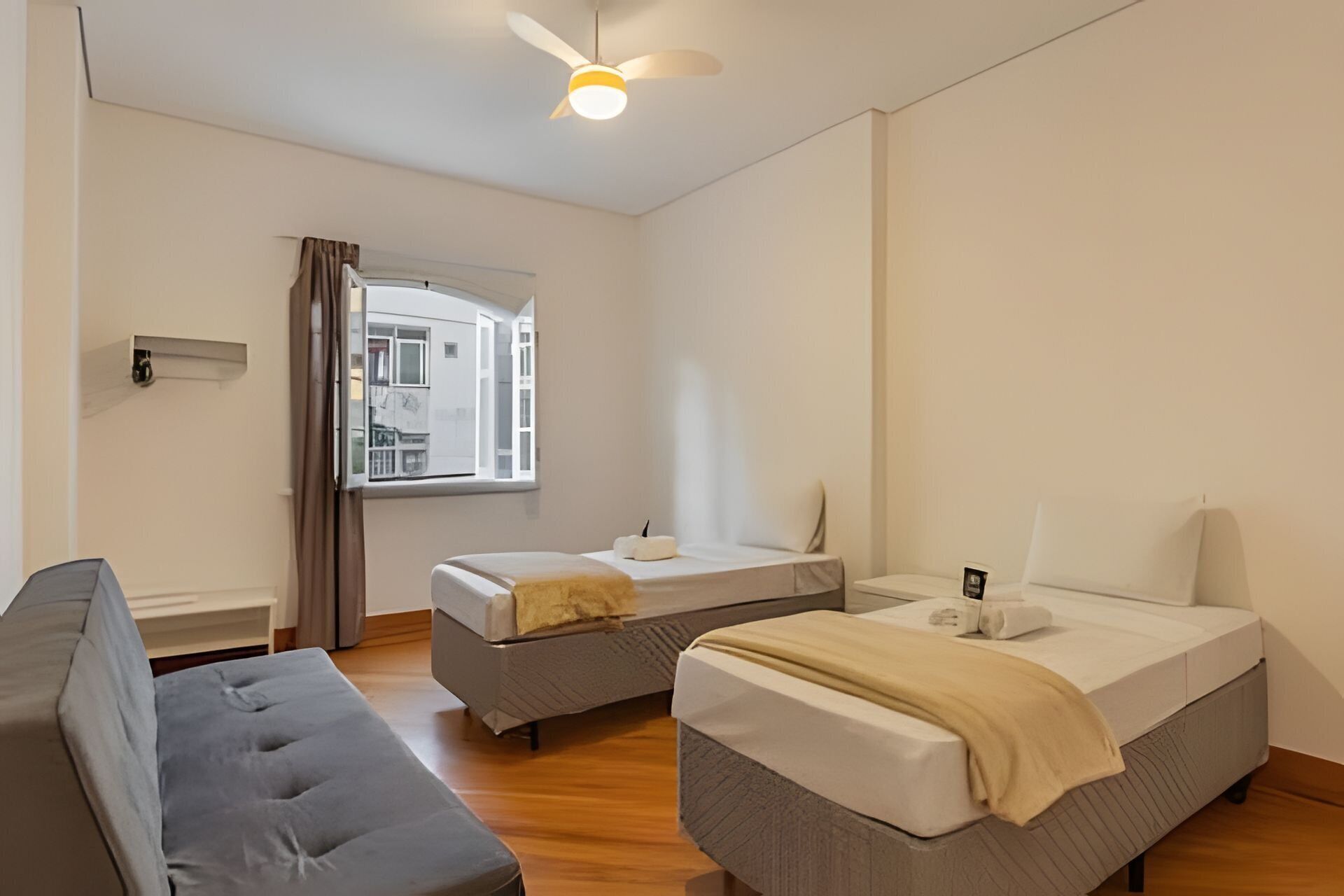 A room with two beds, a sofa, a ceiling fan, and a window with a view of buildings.