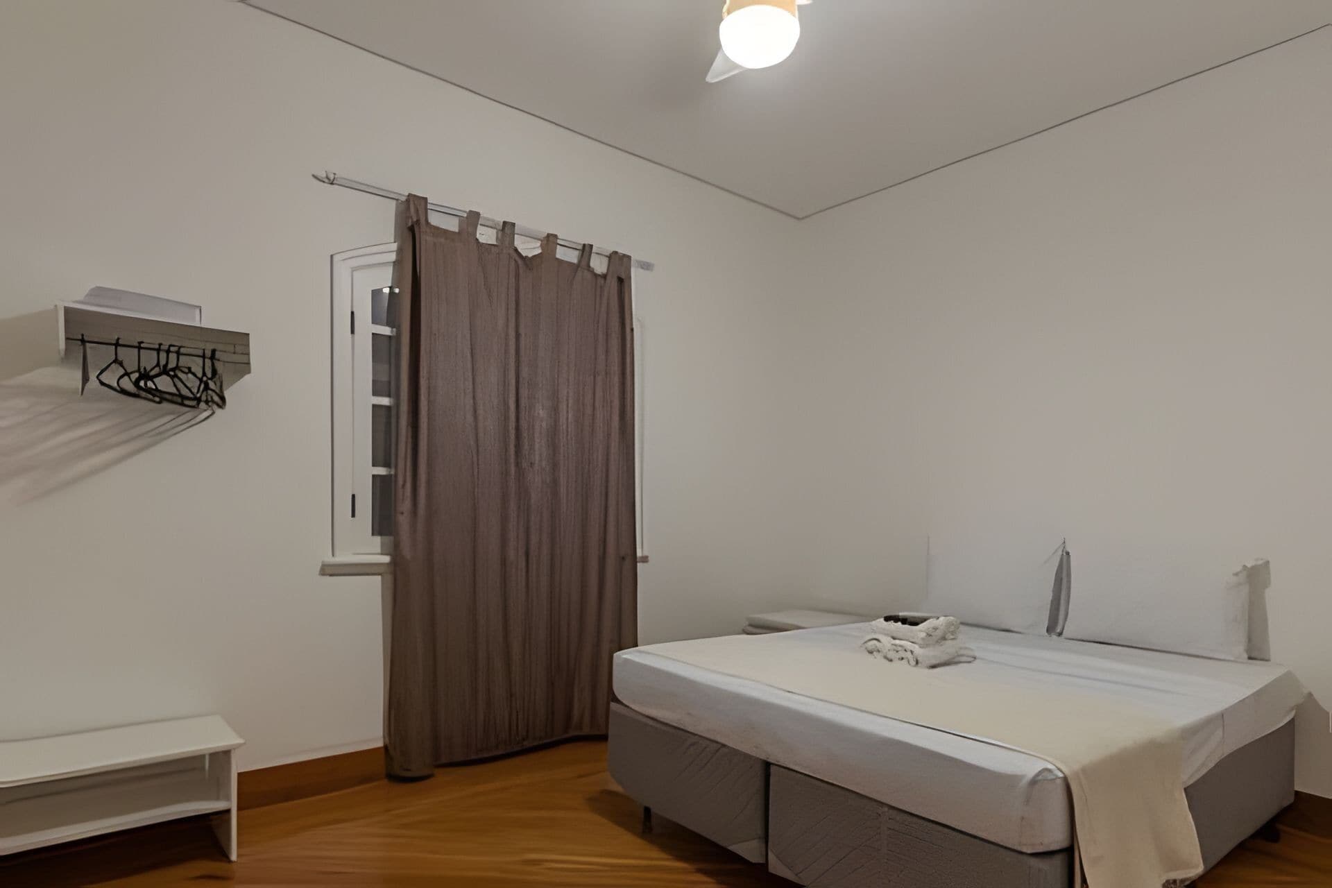 A bedroom with a bed, a window with curtains, a bedside table, and a wall-mounted shelf.
