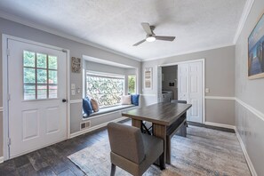 Une pièce comprenant une table à manger, un banc avec des coussins, un ventilateur de plafond et une fenêtre donnant sur des arbres.