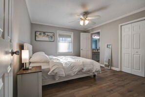 Un dormitorio con cama, mesita de noche, ventilador de techo, ventana con persianas y puerta que da a un baño.