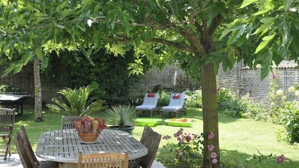 Een terras met een ronde houten tafel en stoelen, een plant in pot, en een kleine tuin met een boom en bloemen.