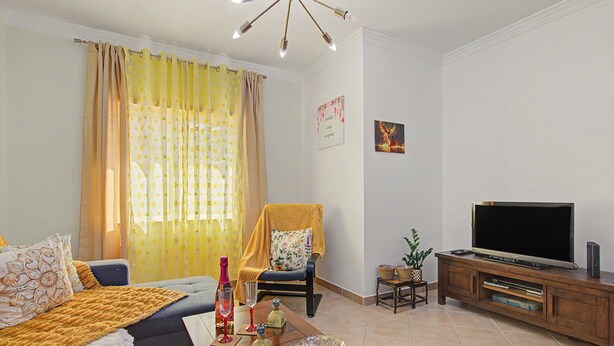 Uma sala de estar com sofá, televisão, aparador de madeira e uma poltrona decorativa.