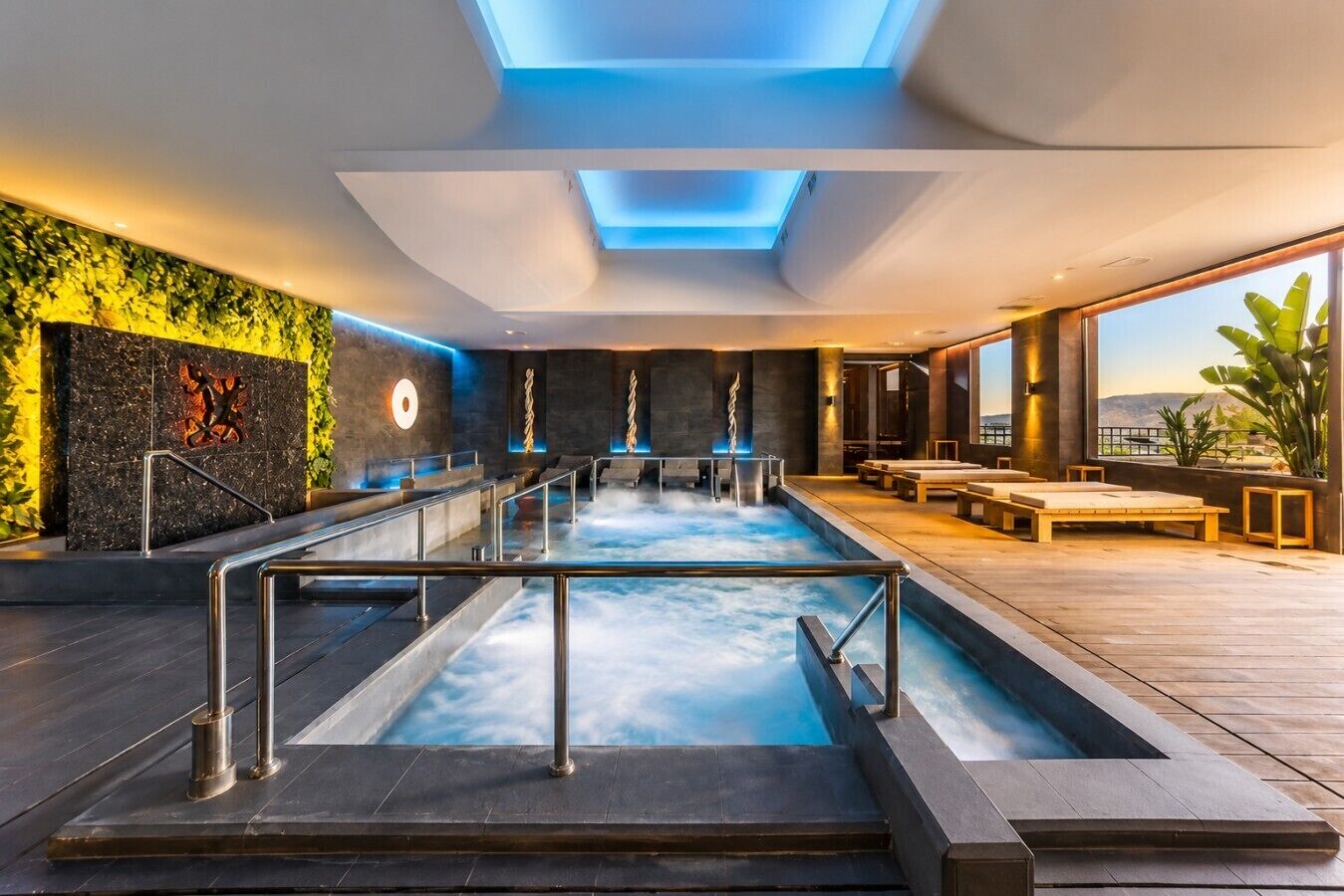 Piscina interior moderna con barrera de vidrio, zona de estar y vistas al exterior.