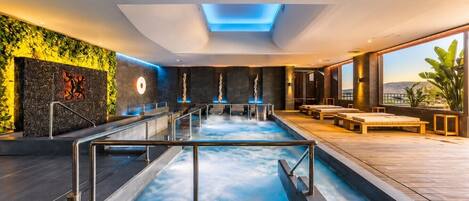 A modern indoor pool with a glass barrier, seating area, and a view of the outdoors.