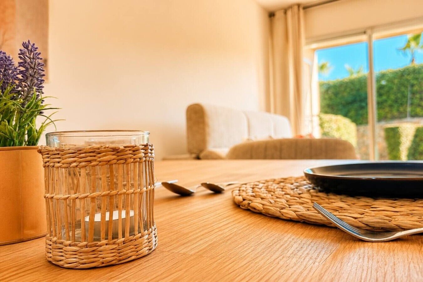 Ein Zimmer mit Holztisch, einem Glasgefäß in einem gewebten Korb, einer Pflanze und Blick auf Grün durch ein Fenster.