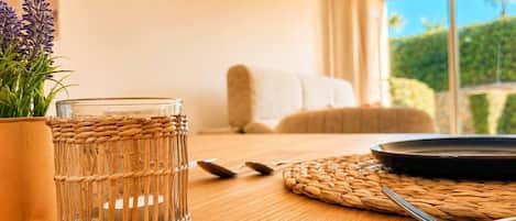 A room with a wooden table, a glass in a woven basket, a plant, and a view of greenery through a window.