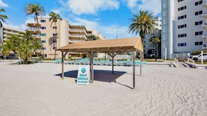Ein Strandverleihbereich mit überdachter Sitzgelegenheit, einem Schild für Strandverleih und Gebäuden im Hintergrund.