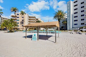 Ein Strandverleihbereich mit überdachter Sitzgelegenheit, einem Schild für Strandverleih und Gebäuden im Hintergrund.