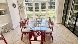 Uma sala de jantar com mesa de vidro, cadeiras vermelhas e vista para um jardim através de grandes janelas.
