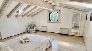Quarto com teto inclinado, uma cama, uma cadeira, uma janela pequena e um espelho.