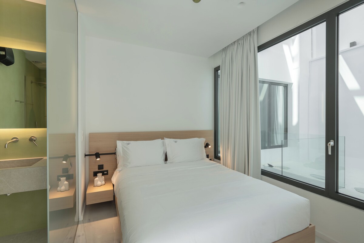 Habitación de hotel moderna con una cama grande, cabina de ducha de vidrio, tocador con espejo y vistas al exterior.