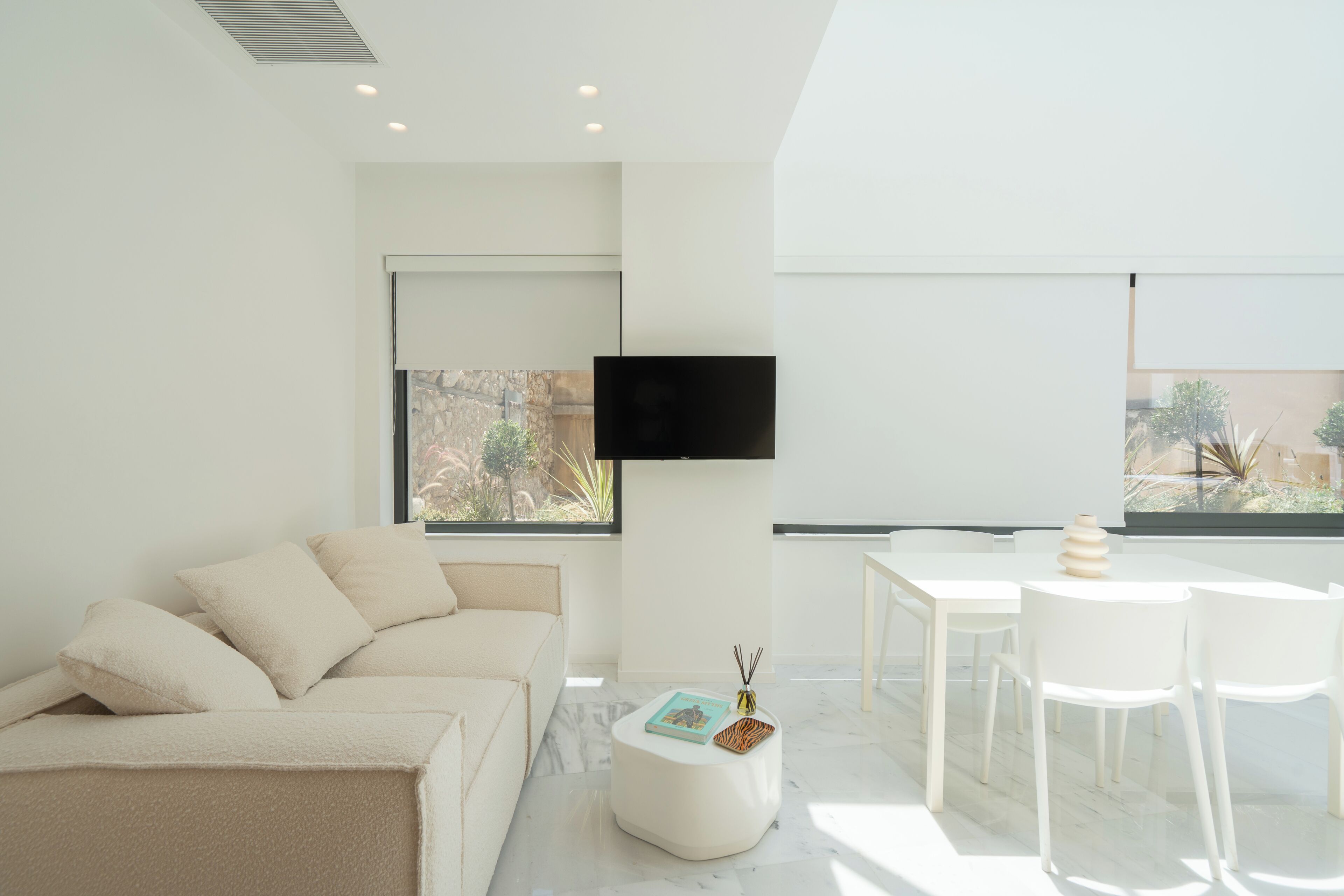 Una sala de estar moderna con un sofá, un televisor de pantalla plana montado en la pared, una zona de comedor con mesa y sillas, y vistas al exterior a través de grandes ventanales.