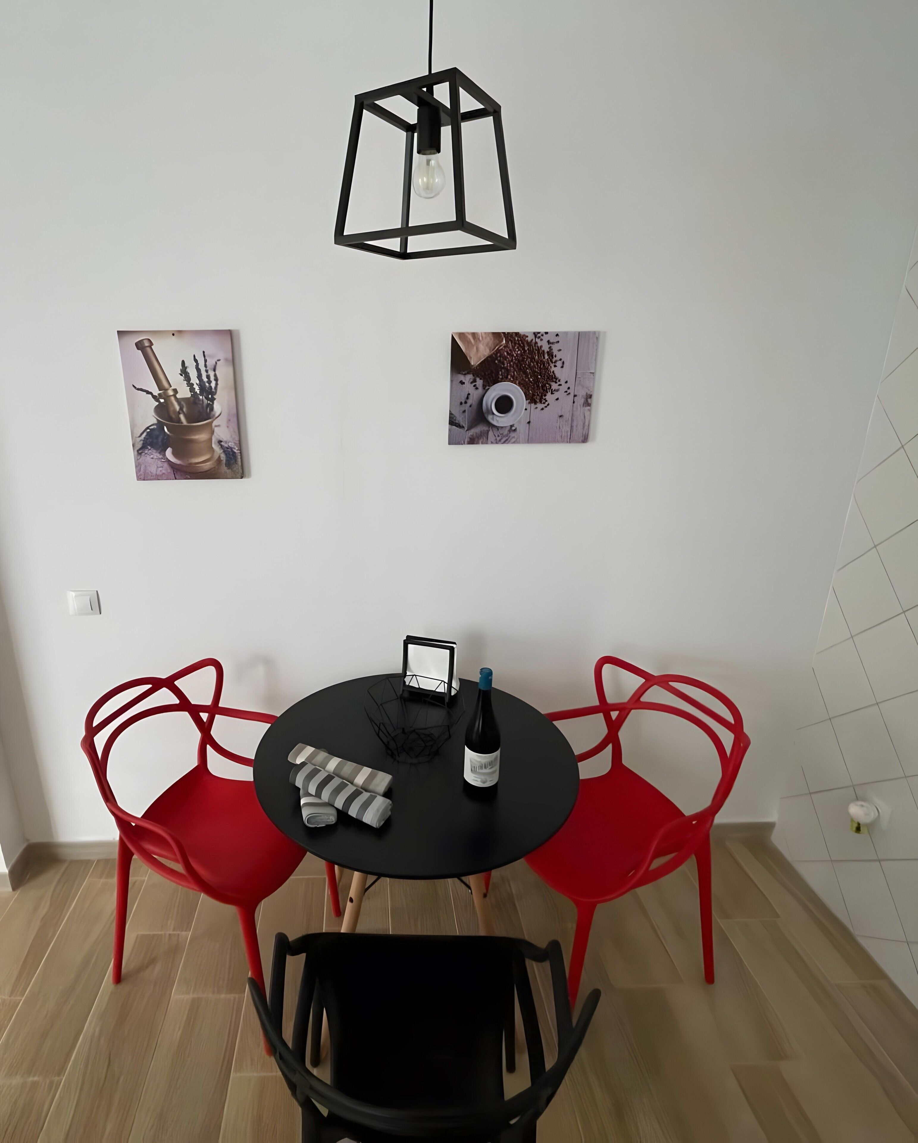 A dining area with a round table, two red chairs, a black chair, a hanging light fixture, and two wall-mounted artworks.
