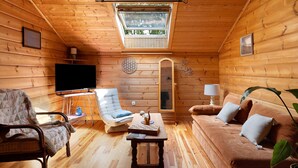 Interior acogedor de una cabaña de troncos con una televisión, un sillón con cojines, un sofá, una mesita y un tragaluz.