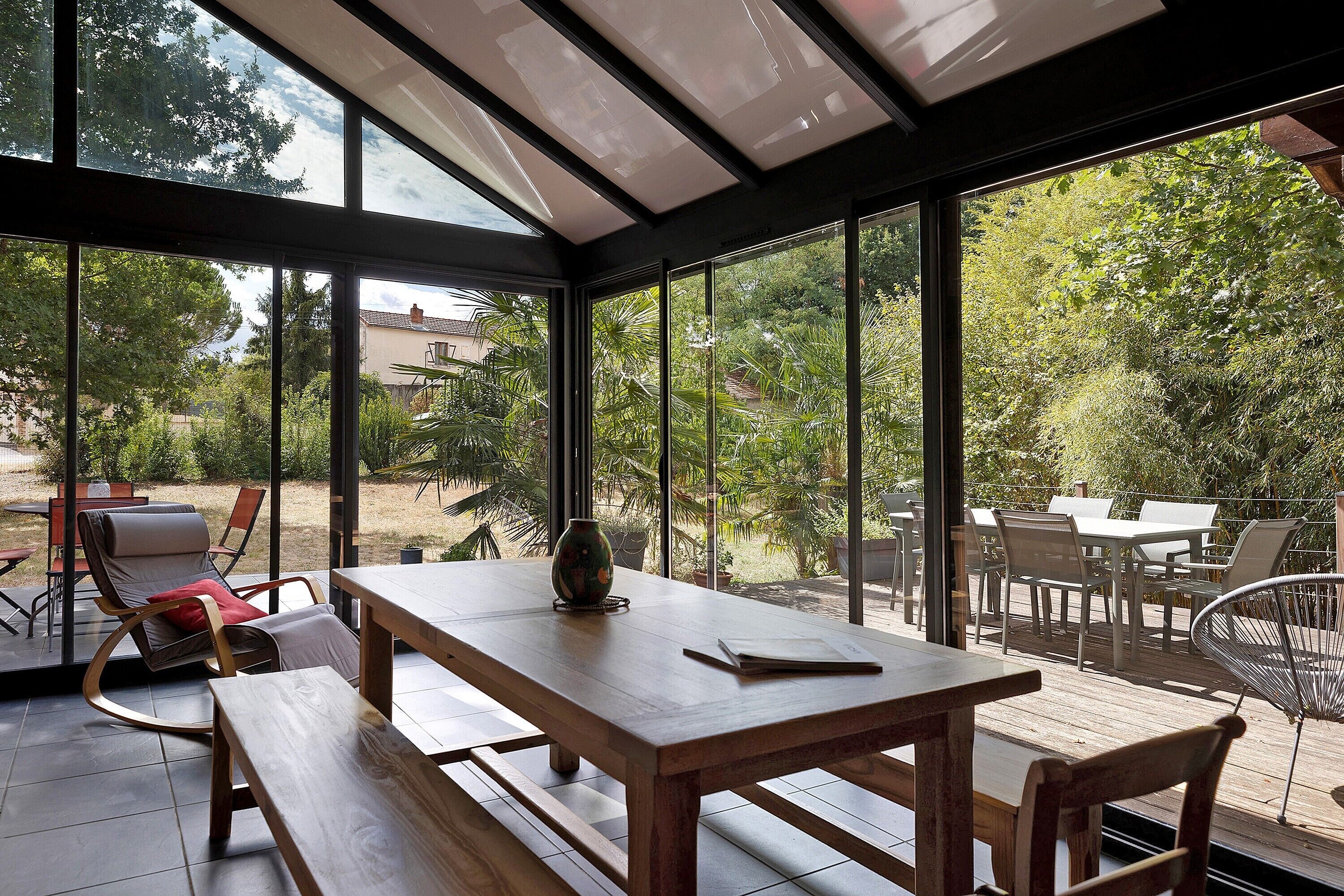 A conservatory with a wooden table and benches, large glass windows, and a view of a garden.