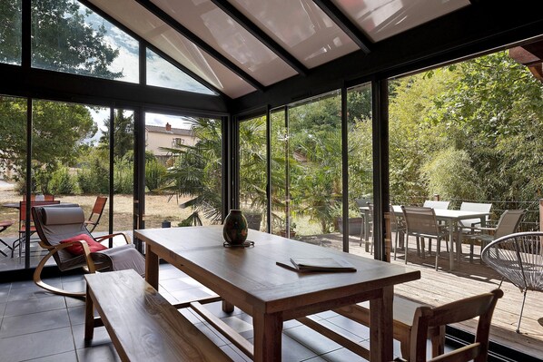 Une véranda avec une table et des bancs en bois, de grandes fenêtres en verre et une vue sur un jardin.