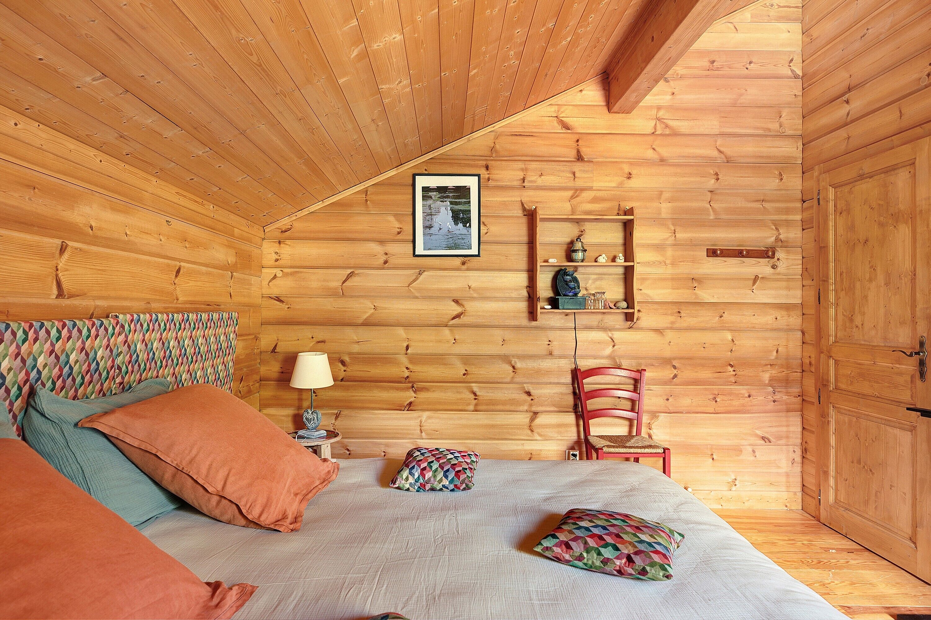A cozy wooden room with a bed, pillows, a chair, a lamp, and a shelf with decorative items.