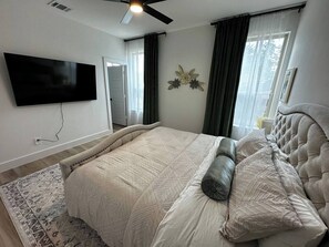 Une chambre à coucher avec un lit, un ventilateur de plafond, une télévision et une fenêtre avec des rideaux.