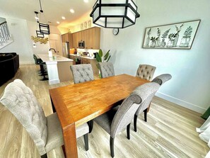 Un coin repas avec une table en bois et des chaises rembourrées, une cuisine à l’arrière-plan et une horloge fixée au mur.