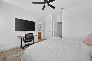 Ein Schlafzimmer mit einem Bett, einem Schreibtisch, einem Stuhl, einem Deckenventilator und einem an der Wand befestigten Fernseher.
