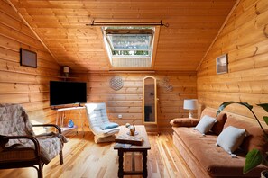 A cozy log cabin interior with a television, a cushioned chair, a sofa, a small table, and a skylight.
